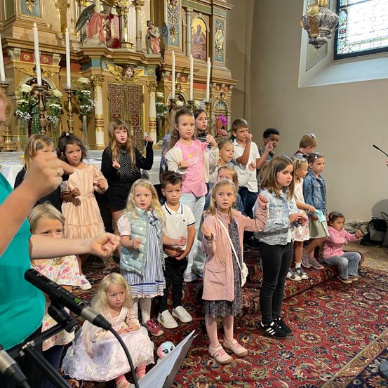 Eine Gruppe von Kindern steht in einer Kirche, einige singen, andere schauen zu. Ein Teppich bedeckt den Boden. Vor ihnen steht ein Mikrofon. Kerzen stehen auf dem Altar.