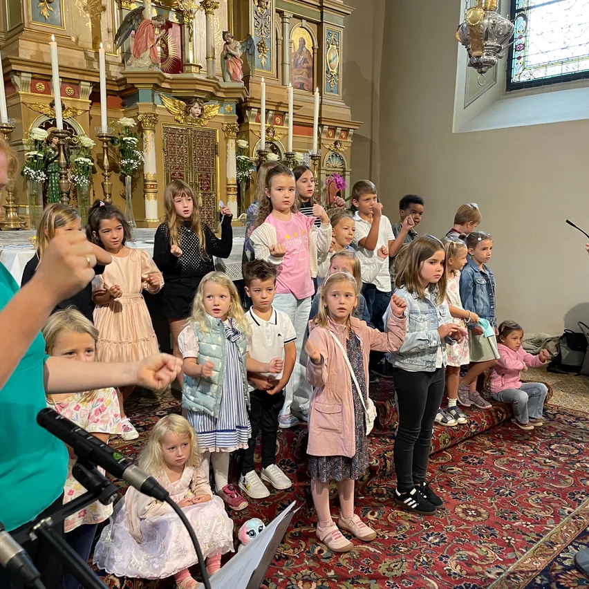 Eine Gruppe von Kindern steht in einer Kirche, einige singen, andere schauen zu. Ein Teppich bedeckt den Boden. Vor ihnen steht ein Mikrofon. Kerzen stehen auf dem Altar.