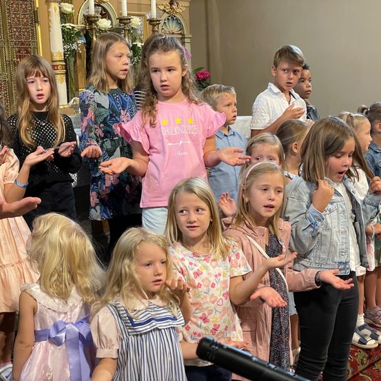 Eine Gruppe von Kindern singt in einer Kirche mit Kerzen und Blumen im Hintergrund. Einige tragen Freizeitkleidung, andere Kleider.