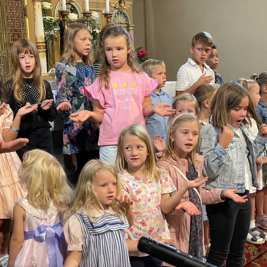 Eine Gruppe von Kindern singt in einer Kirche mit Kerzen und Blumen im Hintergrund. Einige tragen Freizeitkleidung, andere Kleider.