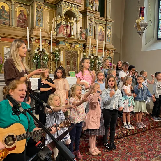 Ein Kinderchor singt in einer Kirche, begleitet von einer erwachsenen Frau, die Gitarre spielt. Die Kinder stehen auf einem Teppich und singen, während die Frau sie anführt. Im Hintergrund befindet sich ein geschmückter Altar mit Kerzen und religiösen Bildern.