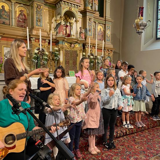 Ein Kinderchor singt in einer Kirche, begleitet von einer erwachsenen Frau, die Gitarre spielt. Die Kinder stehen auf einem Teppich und singen, während die Frau sie anführt. Im Hintergrund befindet sich ein geschmückter Altar mit Kerzen und religiösen Bildern.