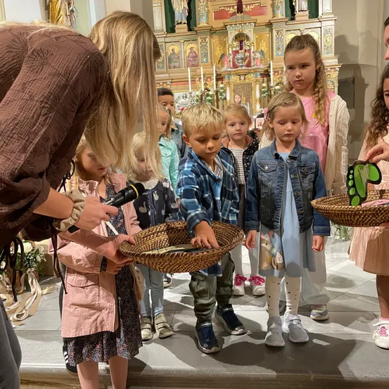 Eine Gruppe von Kindern in einer Kirche, die etwas von einem Erwachsenen mit Mikrofon erhalten. Körbe und dekorative Gegenstände sind vorhanden.