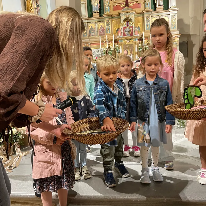 Eine Gruppe von Kindern in einer Kirche, die etwas von einem Erwachsenen mit Mikrofon erhalten. Körbe und dekorative Gegenstände sind vorhanden.