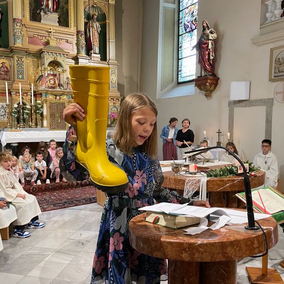 Ein Mädchen hält einen gelben Stiefel in einer Kirche. Viele Kinder sitzen um einen Tisch herum, und Erwachsene stehen in der Nähe. Kerzen und Blumen schmücken den Altar.
