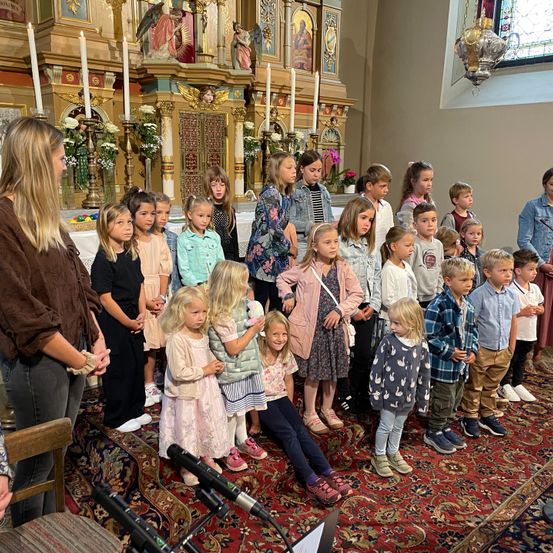 Eine Gruppe von Kindern steht in einer Kirche, wahrscheinlich während einer Zeremonie, mit Erwachsenen in der Nähe. Mikrofone und ein Teppich liegen auf dem Boden.