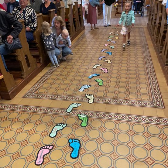 Ein kleines Mädchen läuft einen Kirchengang mit Fußabdrücken auf dem Boden hinunter, Familien und Einzelpersonen sitzen auf Kirchenbänken.
