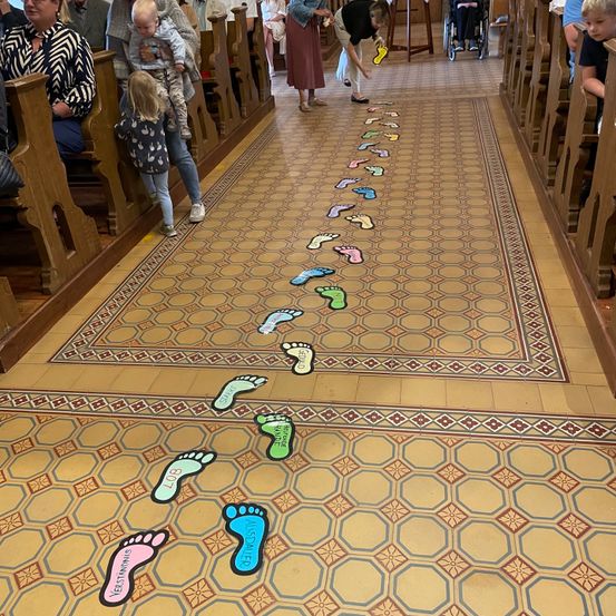 Ein Kirchengang mit dekorativem Fliesenboden, der eine Reihe bunter Fußabdrücke zum Altar hin führt, wo Menschen sitzen und einer steht.