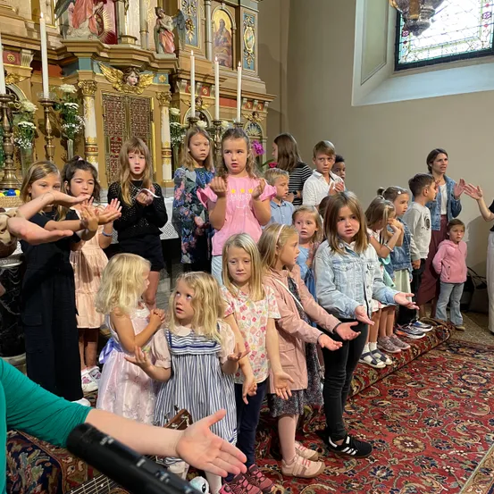 Eine Gruppe von Kindern und Erwachsenen steht mit erhobenen Haenden in einer Kirche, nahe einem Altar mit Kerzen und einem Buntglasfenster.
