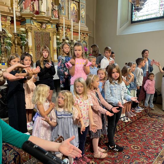 Eine Gruppe von Kindern und Erwachsenen steht mit erhobenen Haenden in einer Kirche, nahe einem Altar mit Kerzen und einem Buntglasfenster.