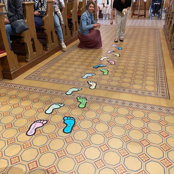 Einige Leute sitzen auf Bänken in einer Kirche. Eine Frau kniet auf dem Boden und hält etwas in der Hand. Der Boden hat Fußabdrücke in verschiedenen Farben.