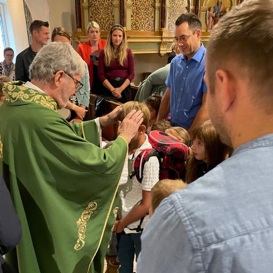 Ein Priester in grünen Gewändern segnet ein Kind in einer Kirche, während andere Menschen um sie herum stehen.