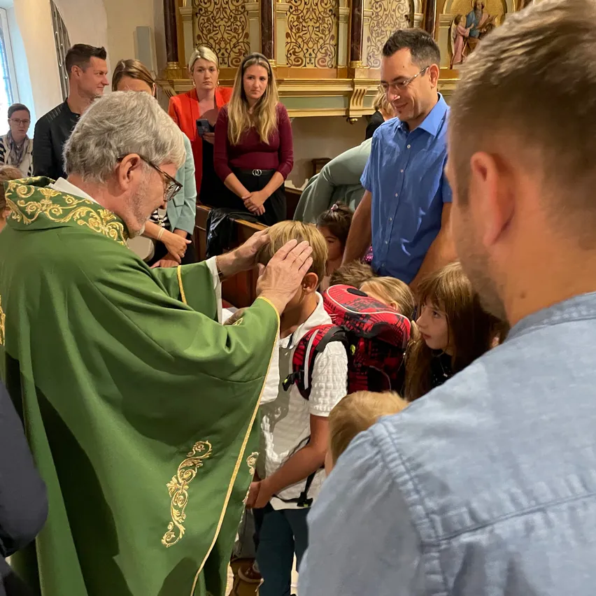 Ein Priester in grünen Gewändern segnet ein Kind in einer Kirche, während andere Menschen um sie herum stehen.