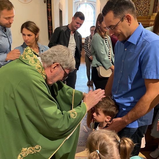 Ein Priester in einer grünen Robe segnet einen kleinen Jungen in einer Kirche, wobei ein Mann und eine Frau seine Arme halten. Andere Menschen stehen im Hintergrund.