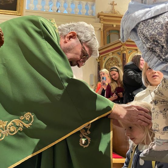 Ein Priester in einer grünen Robe küsst die Hand eines Kindes in einer Kirche. Andere Menschen stehen hinter ihnen.