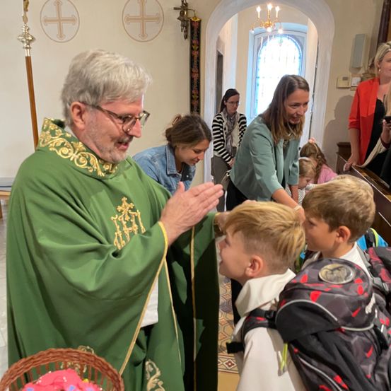 Ein Priester in grüner Amtstracht segnet Kinder in einer Kirche, während Erwachsene zusehen. Kreuze schmücken die Wände, und eine Kronleuchter hängt von der Decke.