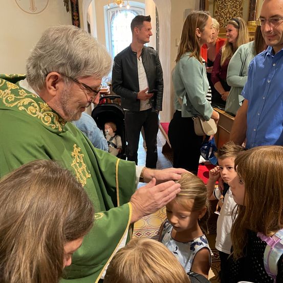 Ein Priester gibt einem jungen Mädchen in einer Kirche einen Segen. Erwachsene und Kinder sind anwesend, einige beobachten. Ein Mann in einer Lederjacke steht im Hintergrund.