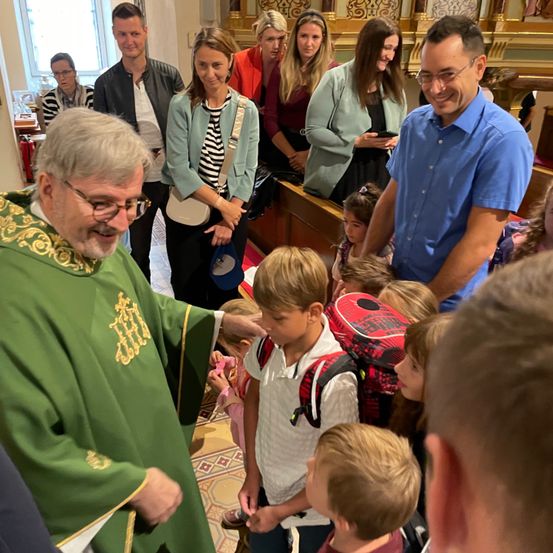 Ein Priester in einer grünen Robe segnet Kinder in einer Kirche, umgeben von Erwachsenen und Kindern, einige mit Rucksäcken.