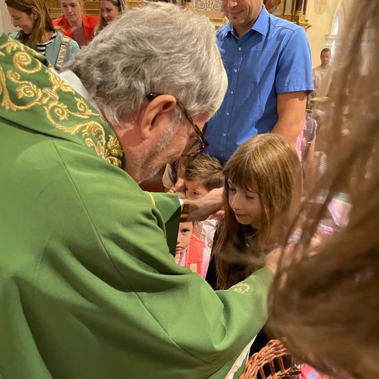Ein Priester in einer grünen Robe berührt die Stirn eines jungen Mädchens in einer Kirche. Andere Menschen, einschließlich eines Mannes in einem blauen Hemd, stehen hinter ihnen.