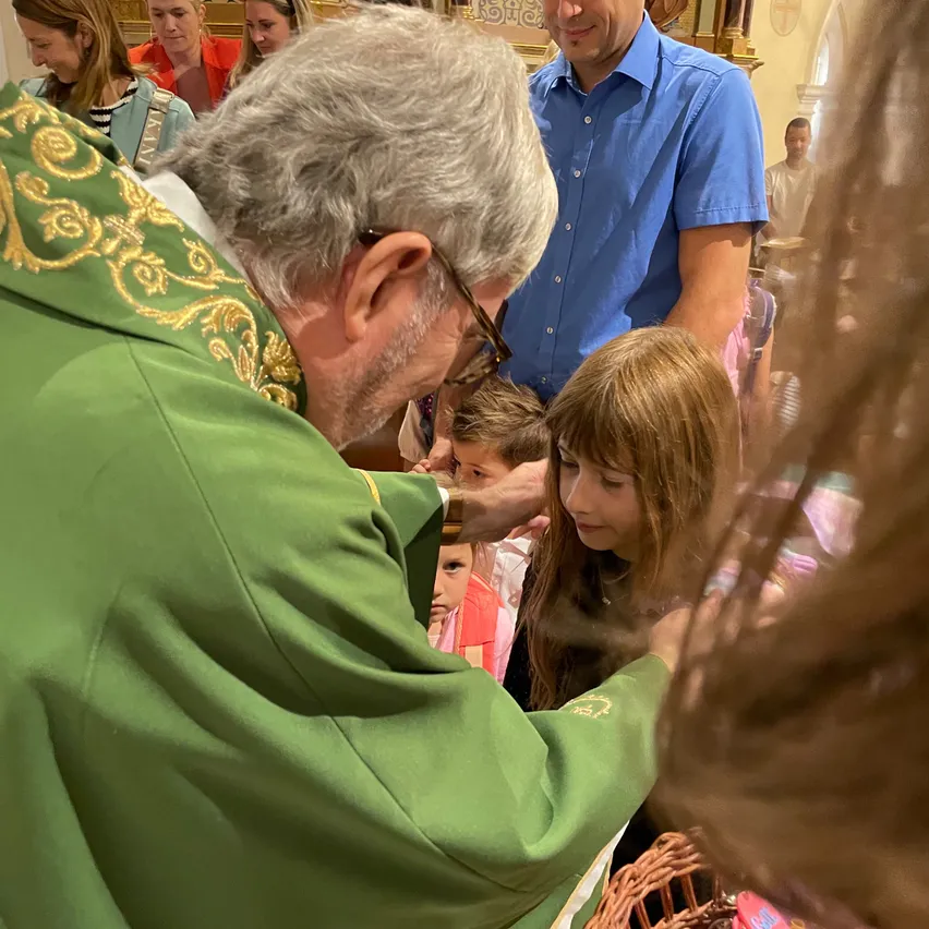 Ein Priester in einer grünen Robe berührt die Stirn eines jungen Mädchens in einer Kirche. Andere Menschen, einschließlich eines Mannes in einem blauen Hemd, stehen hinter ihnen.