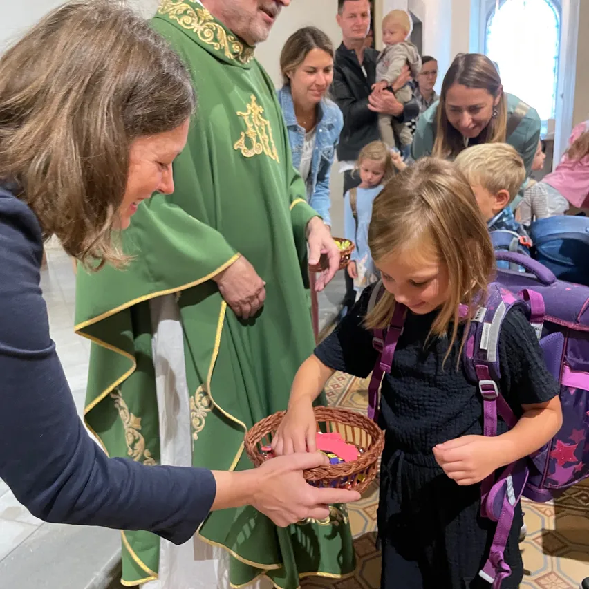 Ein Priester in einer grünen Robe reicht einer jungen Mädchen einen Korb, die einen Rucksack trägt. Das Mädchen schaut auf den Korb, während eine Frau neben ihr lächelt. Andere Menschen sind im Hintergrund, einige tragen Kinder.