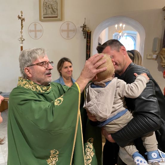 Ein Priester in einer grünen Robe segnet ein Baby, das von einem lächelnden Mann in einer Kirche gehalten wird. Eine Frau steht hinter ihnen.