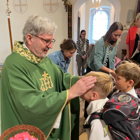 Ein Priester in einem grünen Gewand segnet einen jungen Jungen mit seinen Händen, während andere zusehen. Der Junge trägt einen Rucksack. Hinter ihnen hält eine Frau ein Baby. Der Raum hat eine dekorative Wand und einen gewölbten Eingang.