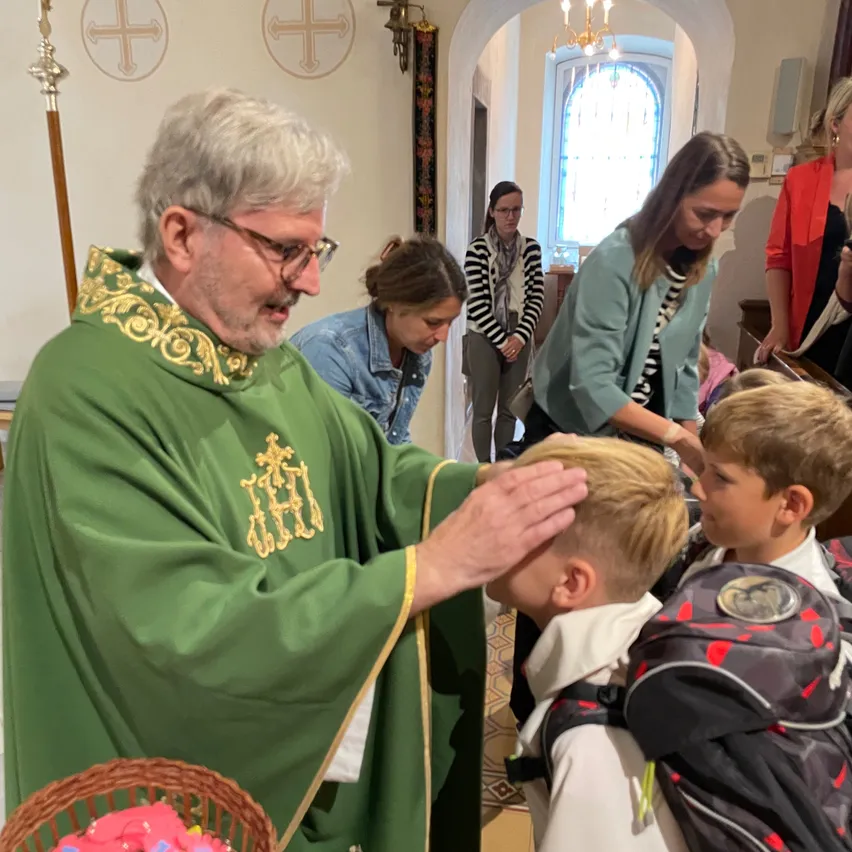Ein Priester in einem grünen Gewand segnet einen jungen Jungen mit seinen Händen, während andere zusehen. Der Junge trägt einen Rucksack. Hinter ihnen hält eine Frau ein Baby. Der Raum hat eine dekorative Wand und einen gewölbten Eingang.