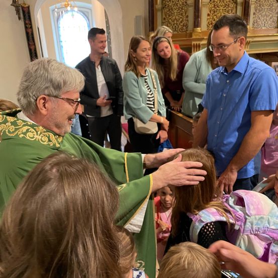 Ein Priester in grünen Roben segnet Kinder in einer Kirche, während Erwachsene zuschauen und stehen.