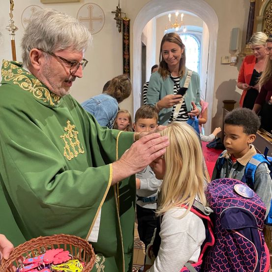 Ein Priester in einer grünen Robe segnet ein junges Mädchen in einer Kirche. Hinter ihnen beobachten eine Frau und Kinder.