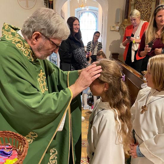 Ein Priester in grünen Gewändern segnet ein junges Mädchen, während andere Menschen in einer Kirche stehen.