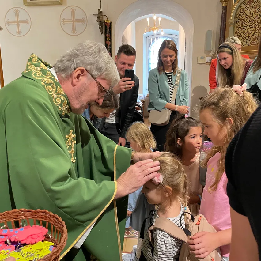 Ein Priester in einem grünen Gewand segnet ein Kind in einer Kirche, während mehrere andere Kinder und Erwachsene zusehen.