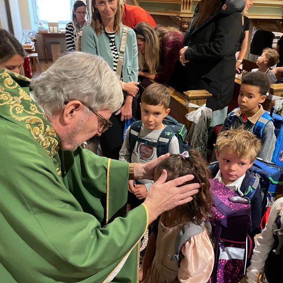 Ein Priester in einem grünen Gewand segnet ein junges Mädchen mit gesenktem Kopf in einer Kirche, umgeben von Kindern und Erwachsenen.