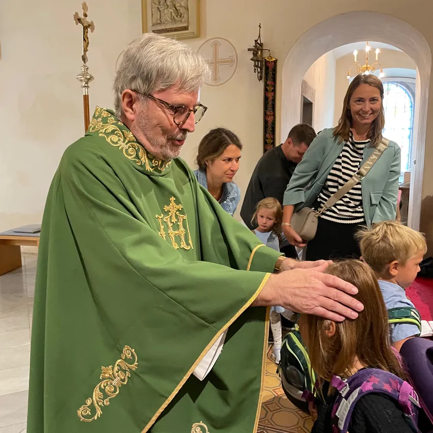 Ein Priester in grüner Amtskleidung segnet ein Kind in einer Kirche, während Familien und Kinder zusehen.