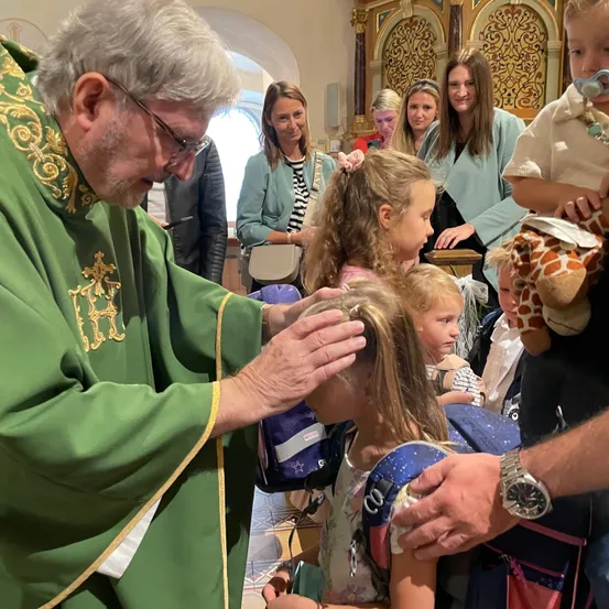 Ein Priester segnet den Kopf eines jungen Mädchens in einer Kirche. Andere Kinder und Erwachsene sind anwesend.