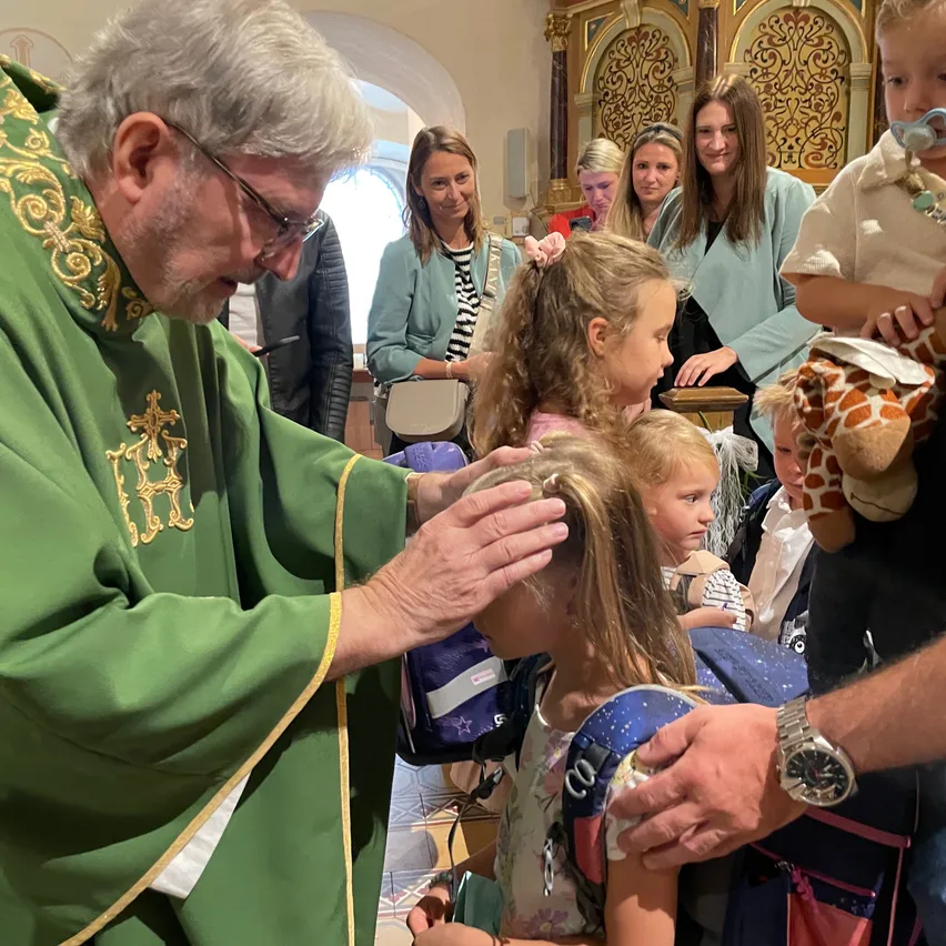 Ein Priester segnet den Kopf eines jungen Mädchens in einer Kirche. Andere Kinder und Erwachsene sind anwesend.