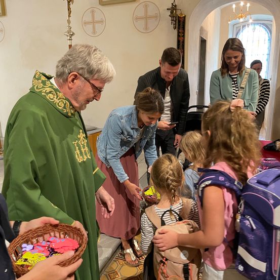 Ein Priester in grünen Gewändern verteilt farbenfrohe Gegenstände an Kinder in einer Kirche, während andere zusehen, darunter eine Frau in einer Jeansjacke und ein Mann in einer schwarzen Lederjacke.