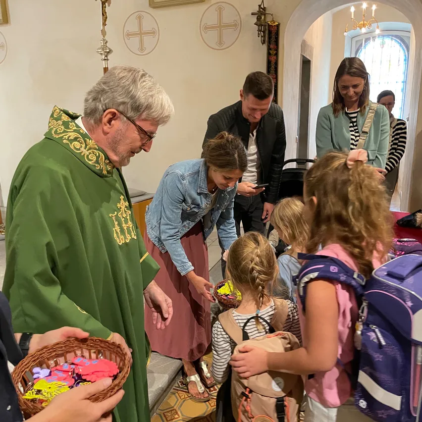 Ein Priester in grünen Gewändern verteilt farbenfrohe Gegenstände an Kinder in einer Kirche, während andere zusehen, darunter eine Frau in einer Jeansjacke und ein Mann in einer schwarzen Lederjacke.