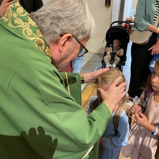 Ein Priester in einer grünen Robe segnet ein junges Mädchen, während andere, darunter ein Baby in einem Kinderwagen, zusehen.