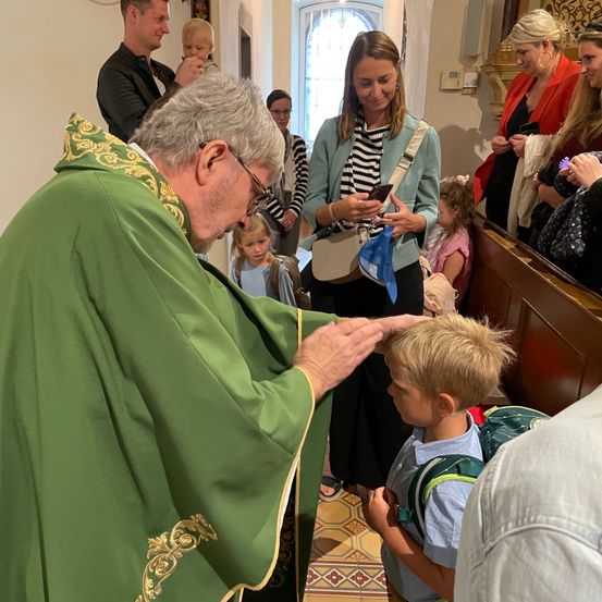 Ein Priester in einem grünen Gewand segnet einen jungen Jungen mit einem Rucksack, während eine Frau ein Kind hält und andere im Inneren einer Kirche zusehen.