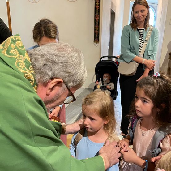 Ein Priester in grünen Gewändern segnet zwei junge Mädchen, eines trägt einen Rucksack. Hinter ihnen steht eine Frau mit einem Kinderwagen.