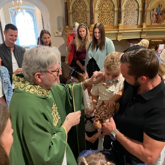 Ein Priester in grünen Gewändern segnet ein Kind in einer Kirche, umgeben von Menschen, die Fotos machen und zusehen.
