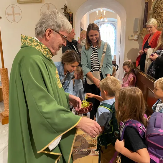 Ein Priester in einer grünen Robe gibt Kindern in einer Kirche Geschenke. Einige Erwachsene und Kinder schauen zu.