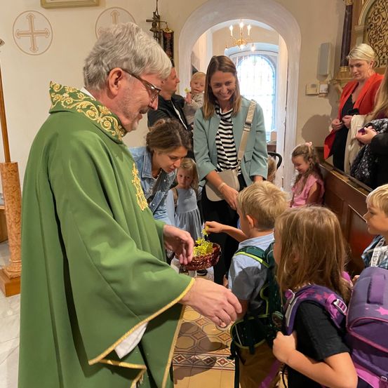 Ein Priester in einer grünen Robe gibt Kindern in einer Kirche Geschenke. Einige Erwachsene und Kinder schauen zu.