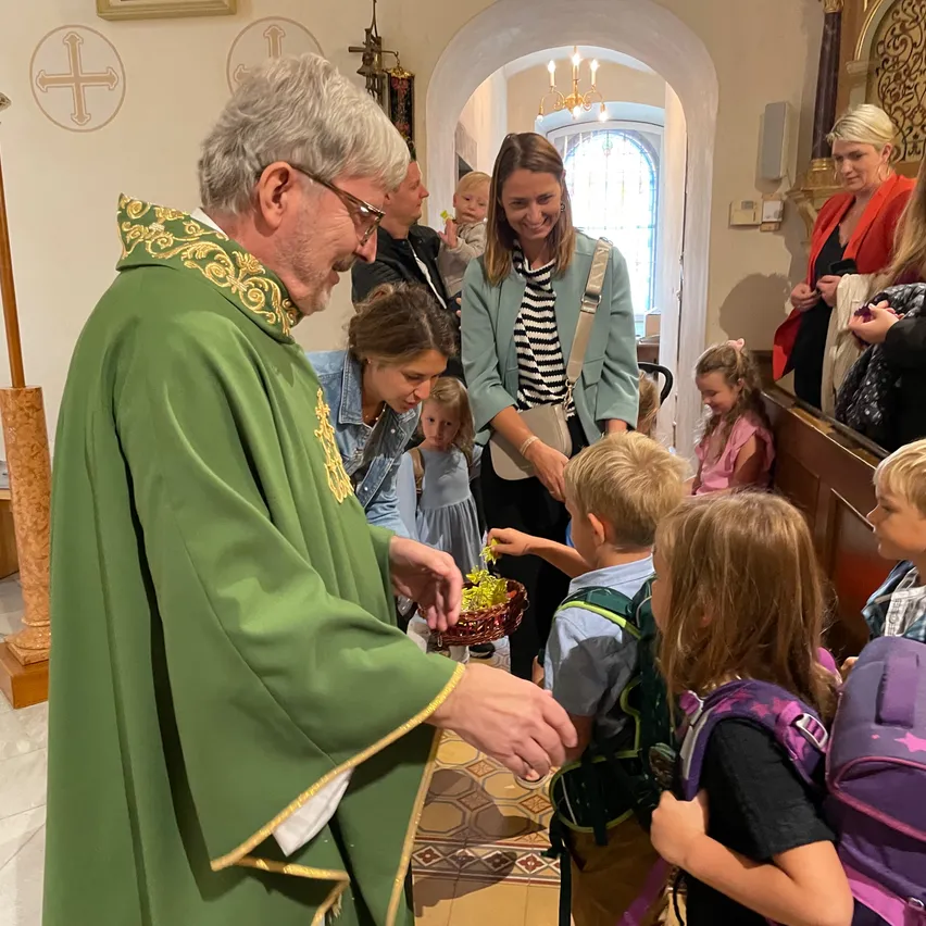 Ein Priester in einer grünen Robe gibt Kindern in einer Kirche Geschenke. Einige Erwachsene und Kinder schauen zu.
