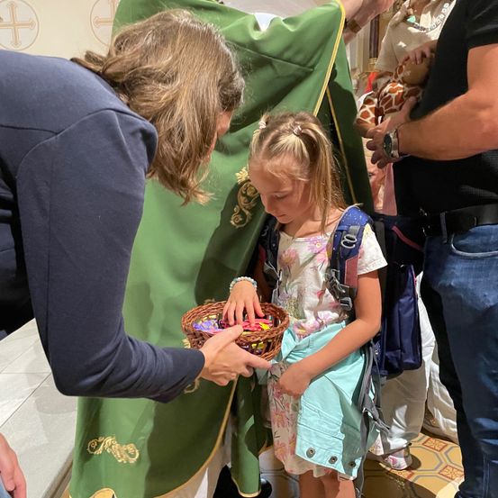 Ein kleines Mädchen mit einem Korb voller Schokoladen steht vor einer Person in einer grünen Robe. Hinter ihnen hält eine andere Person eine Babypuppe. Die Szene spielt sich in einem Raum mit einem gemusterten Boden ab.