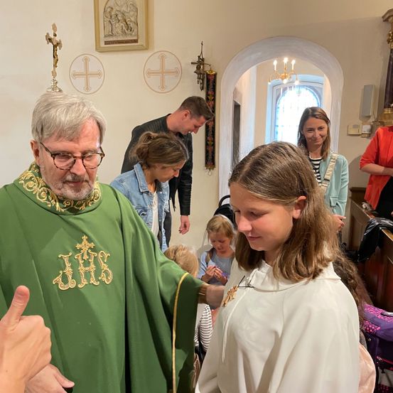 Ein Priester in grüner Robe segnet ein Mädchen in einer weißen Robe. Andere Personen stehen und beobachten. Die Umgebung ist in Innenräumen, mit religiösen Symbolen an der Wand.