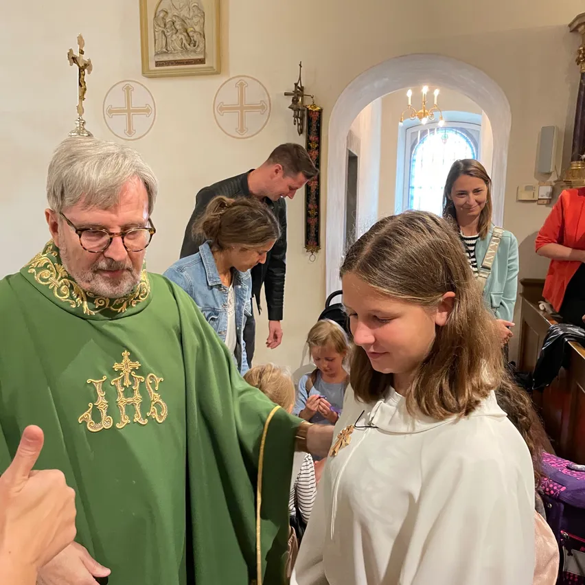 Ein Priester in grüner Robe segnet ein Mädchen in einer weißen Robe. Andere Personen stehen und beobachten. Die Umgebung ist in Innenräumen, mit religiösen Symbolen an der Wand.
