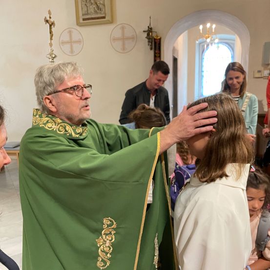 Ein Priester in einer grünen Robe segnet ein Mädchen in einer Kirche. Dahinter beobachten ein Mann und eine Frau. Eine Kronleuchter und religiöse Symbole schmücken die Wand.