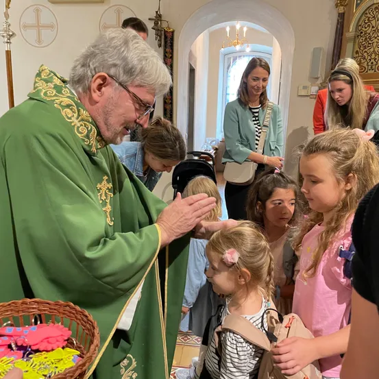 Ein Priester in einer grünen Robe segnet eine Gruppe junger Mädchen in einer Kirche, von denen eine ein Korb mit bunten Schlüsselanhängern hält.